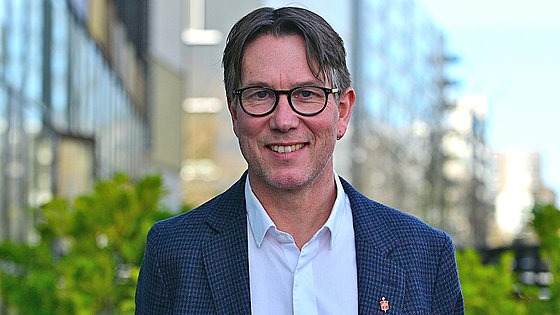 Ein Mann, dunkle Haare mit Mittelscheitel, links ein Ohrring, Brille, weißes offenes Hemd, dunkles Jackett mit Bischofsnadel, blickt freundlich lächelnd in die Kamera. Im Hintergrund Häuser und grüne Büsche.