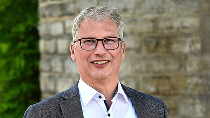 Das Bild zeigt ein Porträt eines Mannes mittleren Alters mit grau melierten, kurzen Haaren. Er trägt eine Brille und ein graues Sakko über einem weißen Hemd mit Knöpfen. Der Hintergrund ist unscharf und zeigt eine helle Steinmauer.