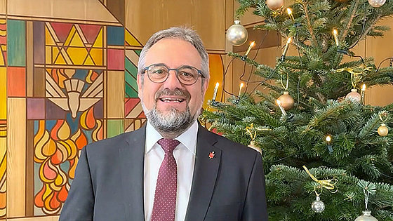 Ein Mann im schwarzen Jackett mit weißem Hemd und roter Krawatte. Rechts hinter ihm ist ein geschmückter Weihnachtsbaum. Links hinter ihm ist die bunt gestaltete Stirnwand eines Gottesdienstraums zu sehen.
