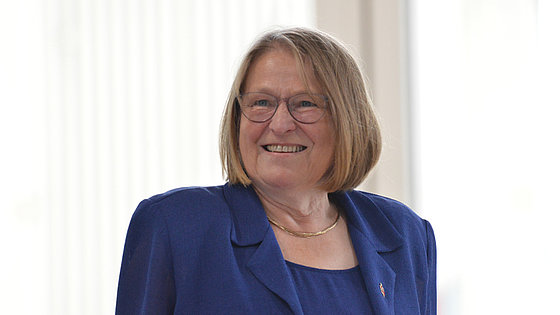 Eine Frau, dunkelblondes Haar, Brille, Kette, blaues Kostüm mit Bischofsnadel am Revers, lächelt freundlich vor sehr hellem Hintergrund.