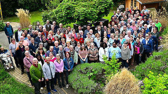 Das Bild zeigt die Konferenzmitglieder, die für ein Gruppenfoto posieren. Sie stehen im Freien, vor einem Garten mit Büschen und Bäumen. Im Hintergrund ist ein kleines Gartenhaus zu sehen. Die Menschen sind unterschiedlich alt und tragen verschiedene Kleidung.
