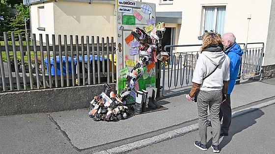 Zwei Personen stehen auf einer Straße und betrachten eine alte Tür, die an einem Holzzaun befestigt ist. Vor der Tür ist schräg ein Kreuz aus Schuhen festgemacht. Auf der Tür und auf dem Kreuz kleben Zettel mit Botschaften zum Nachdenken.