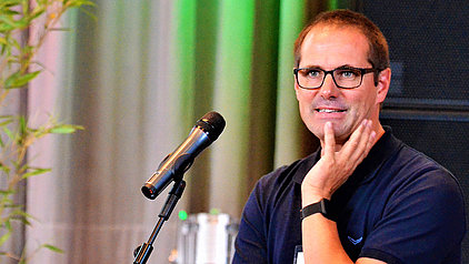 Das Bild zeigt einen Mann mittleren Alters mit kurzen, braunen Haaren und einer Brille. Er trägt ein dunkelblaues Poloshirt und hält seine Hand am Kinn. Er steht vor einem Mikrofon. Im Hintergrund sind Pflanzen und Teile einer Bühne zu sehen.