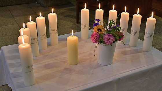 Ein weißer Tisch mit zwölf brennenden weißen Kerzen, die in zwei Reihen angeordnet sind. In der Mitte des Tisches steht eine Vase mit einem bunten Blumenstrauß und einer weiteren Kerze.