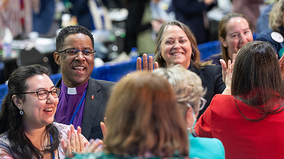 Vier Freude ausstrahlende Gesichter. Bei der Generalkonferenz Anwesende klatschen einander ab. Mit dabei und hier mit im Bild ist Bischof Cedrick Bridgeforth im Kollarhemd und mit Brille.