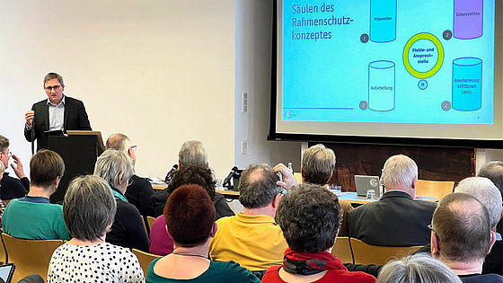 Ein Mann mittleren Alters steht an einem Rednerpult. Die Projektion auf einer großen Leinwand zeigt die Säulen des Rahmenschutzkonzeptes: Prävention, Intervention, Aufarbeitung und Anerkennung des Leids. Das Foto zeigt zudem einen Teil der Delegierten.
