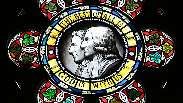 Ein rundes Kirchenfenster mit einem zentralen Medaillon, das die Portraits von John (1703-1791) und Charles (1707-1788) Wesley zeigt, umgeben von einem Band mit der Aufschrift "Das Beste von allem ist, Gott ist mit uns". Das Fenster ist mit floralen Motiven verziert.
