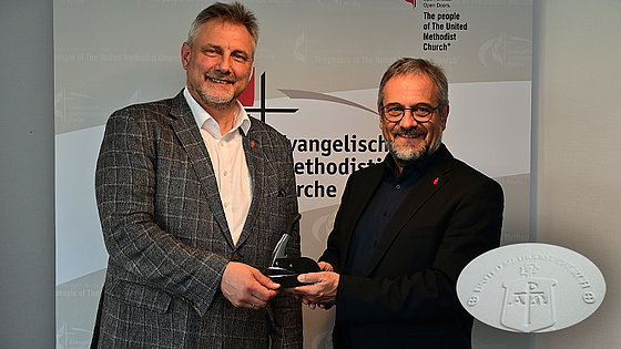 Links ein Mann mit offenem weißem Hemd und kariertem Jackett. Rechts ein Mann mit Brille in offenem dunklem Hemd und schwarzem Jackett. Beide, mit graumeliertem Haar und Bart sowie Bischofsnadel am Revers, lächeln in die Kamera.