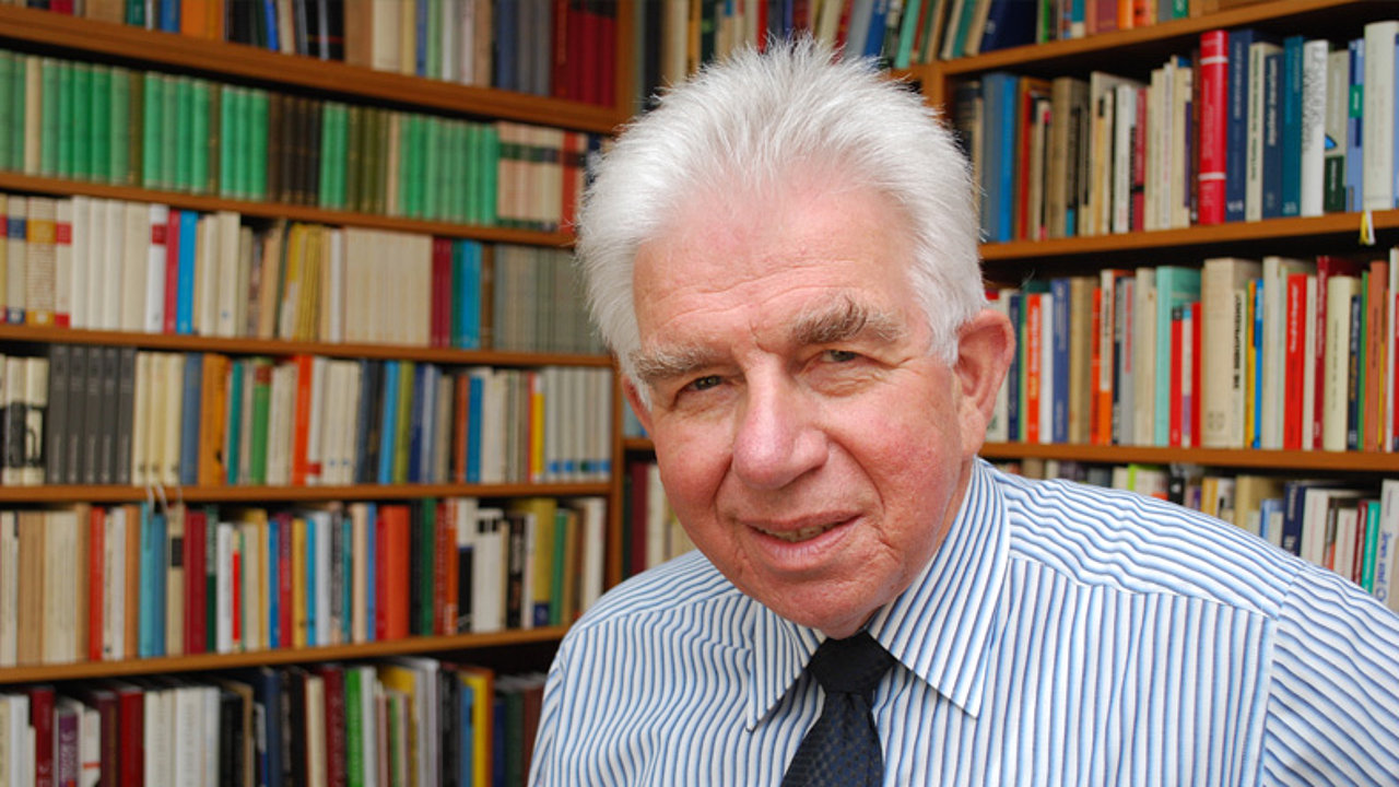 Ein älterer Mann mit weißem Haar, einem gestreiften Hemd und einer Krawatte lächelt in die Kamera. Im Hintergrund sind Regale voller Bücher zu sehen.