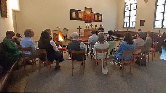 Rund ein Dutzend meist ältere Menschen sitzen auf Stühlen in einem Kirchensaal. Ihre Blicke sind auf eine Frau gerichtet, die sich im erhöhten vorderen Teil des Saals befindet. Auf dem Abendmahlstisch brennen Kerzen. Sonnenlicht erhellt den Raum.