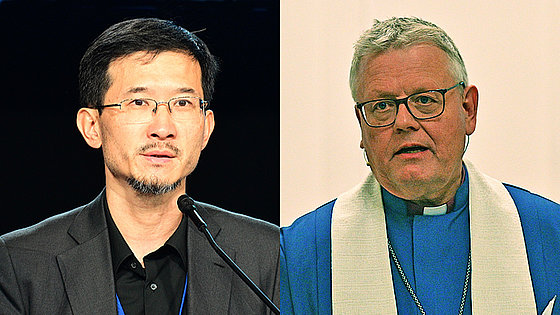 Links: Ein Mann mittleren Alters mit schwarzen Haaren, Oberlippen- und Kinnbart, konzentrierter Blick, dunkles Jackett und schwarzes Hemd, spricht in ein Mikrofon. Rechts ein Mann mit grauem Haar, blauem Bischofsgewand und weißer Stola.