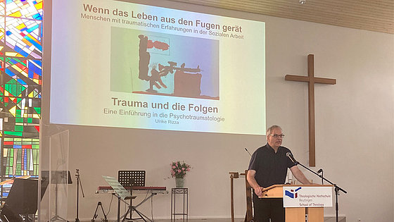 Gottesdienstraum mit Kreuz an der Front. Links davon ein projiziertes Vortragsbild zum Thema Trauma: Ein Strichmännchen, hinterrücks von einer schwarzen Figur bedrängt. Im Vordergrund ein Vortragspult, an dem der Rektor der THR den Traumafachtag eröffnet.
