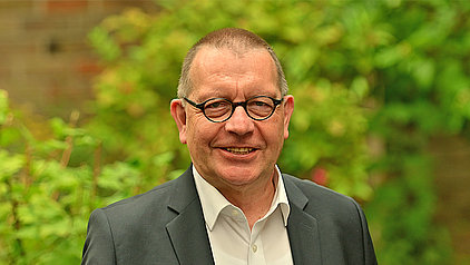 Ein Mann mittleren Alters mit Brille und kurzem, dunklem Haar lächelt freundlich in die Kamera. Er trägt ein weißes Hemd unter einem dunklen Anzugsakko. Der Hintergrund ist unscharf und zeigt grüne Blätter und Pflanzen.