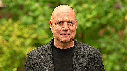 Ein Mann mit Glatze, bekleidet mit einer dunklen Jacke über einem schwarzen T-Shirt, blickt lächelnd in die Kamera. Der Hintergrund ist unscharf und zeigt grünes Blattwerk.