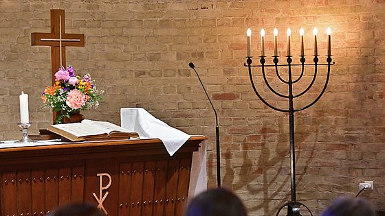 Links der Abendmahlstisch in dunklem Braun, darauf ein Kreuz und davor Blumen und Bibel. Links neben der Bibel eine weiße brennende Kerze. Rechts neben der Bibel Brot und Kelch zugedeckt. Rechts steht die Menora. Im Hintergrund eine Backsteinmauer.