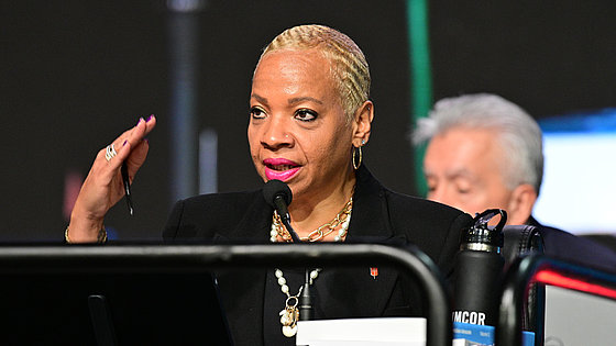 Bischöfin Malone: Afroamerikanerin mit kurzen gebleichten Haaren, Ohr- und Nasenpiercing, Lippenstift, im schwarzen Blazer, spricht in ein Mikrofon. In der rechten Hand hält sie einen Stift und gestikuliert. Im Vordergrund ist ein schwarzes Geländer.