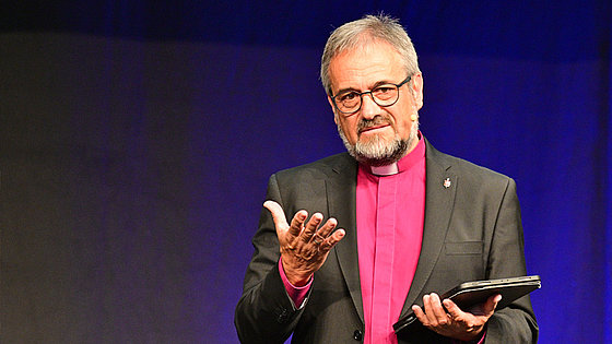Vor einem dunklen Hintergrund steht Bischof Harald Rückert und predigt. Er trägt ein schwarzes Jackett und ein violettes, pastorales Collarhemd. Er streckt seine rechte, geöffnete Hand zum Publikum aus. Sein Blick ist nachdenklich-fragend.