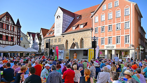Bei wolkenlosem Himmel sitzen hunderte Menschen an Biertischgarnituren auf einem Platz im Schatten. Auf den Tischen stehen Blumen, Getränkeflaschen und Plastikschüsseln. Auf einem Anhänger steht eine Frau und spricht in ein Mikrofon.