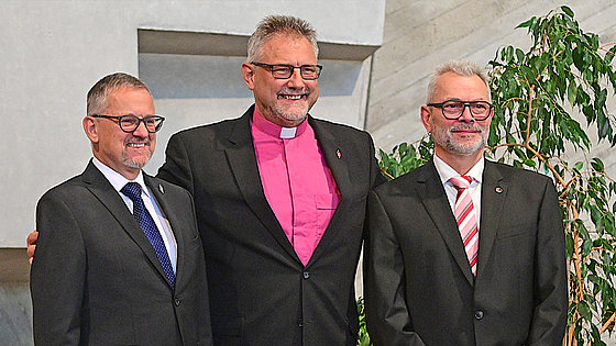 Drei Herren im schwarzen Anzug stehen vor einer Betonwand und einer Pflanze. Der Mittlere trägt ein Kollarhemd, die anderen beiden ein weißes Hemd mit Krawatte. Alle drei lächeln.