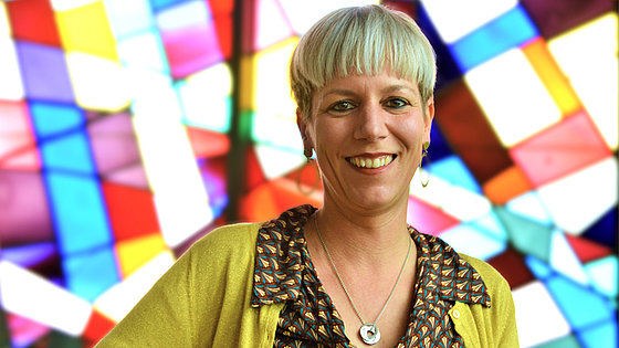 Vor einem Buntglasfenster steht eine fröhlich lächelnde Frau mit heller Kurzhaarfrisur. Sie trägt eine bunte Bluse und senfgelbe Strickjacke. Es ist Pastorin Damaris Hecker, die EmK-Beauftragte für Menschen verschiedener Lebens- und Liebensweisen.