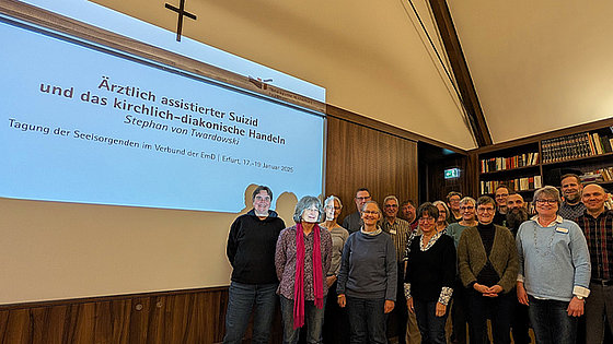 Links ist die erste Seite des Vortrags an die Wand projiziert. Titel: »Ärztlich assistierter Suizid und das kirchlich-diakonische Handeln« von Stephan von Twardowski. Rechts stehen Menschen, die in die Kamera lächeln.