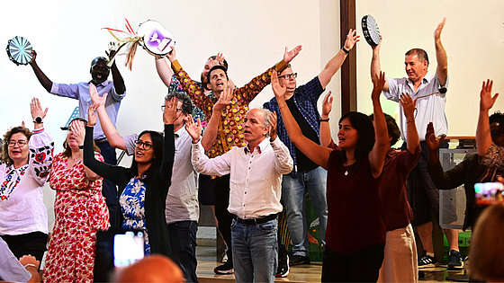 Ein Dutzend Männer und Frauen unterschiedlicher Nationalität singen stehend und mit erhobenen Händen. Einige haben eine Handtrommel in der Hand und begleiten den Gesang.
