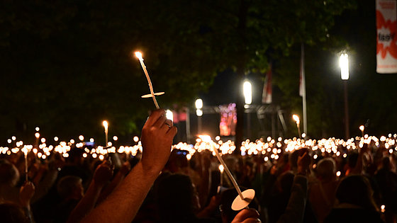 Im Vordergrund zwei dünne brennende Kerzen mit rundem Schutz gegen herunterfließendes heißes Wachs. Im Hintergrund viele weitere brennende Kerzen und Lampen sowie Bäume in der Dunkelheit des Abends.