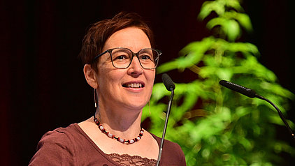 Portrait von Dorothea Lorenz. Eine Frau mit kurzen, rotbraunen Haaren und einer Brille. Sie trägt ein weinrotes Oberteil und eine Perlenkette. Sie lächelt und spricht in ein Mikrofon. Im Hintergrund ist eine unscharfe grüne Pflanze zu sehen.