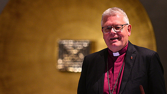 Ein Mann mit grauem Haar, Brille, purpurfarbenem Kollarhemd, schwarzem Jackett mit Bischofsabzeichen im Revers und Brustkreuz spricht in ein Mikrofon.