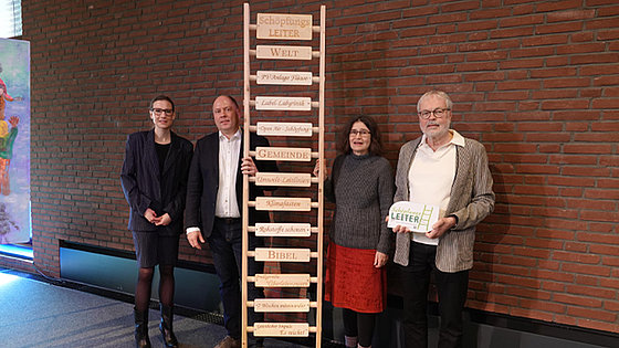 Vor einer roten Ziegelmauer stehen vier Personen. Die mittleren Zwei halten die über zwei Meter hohe Schöpfungs-Leiter aus Holz. An deren Sprossen sind Bretter befestigt, auf denen Begriffe stehen. Die Buchstaben sind eingebrannt.