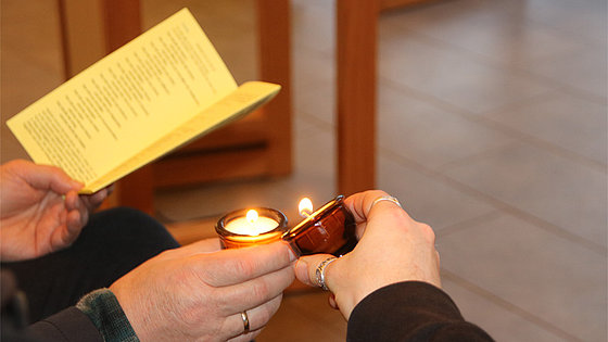 Von zwei Personen sind drei Hände auf dem Foto festgehalten. Die linke Person hält in ihrer Linken ein bedrucktes Blatt Papier und in ihrer Rechten waagrecht ein brennendes Teelicht. Die rechte Person hält in ihrer Rechten ein gekipptes, brennendes Teelicht.