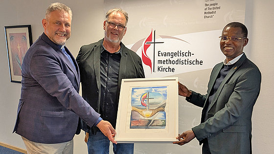 Im Flur der Kirchenkanzlei halten drei Herren in Jacketts gemeinsam ein gerahmtes Gemälde. Es zeigt Kreuz und Flamme sowie das Kreuz in der Weltkugel über zwei offenen Händen, durch die helles Licht scheint. Die unten dargestellte Welt ist zerrissen.