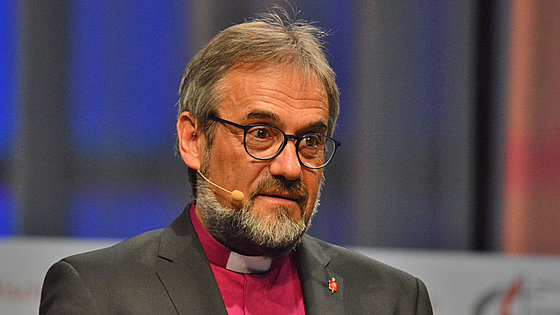 Bischof Rückert, im schwarzen Anzug und violetten Kollarhemd, spricht bei einer Konferenz-Predigt nachdenklich-ernsthaft, mit gerunzelter Stirn zu seiner Zuhörerschaft.