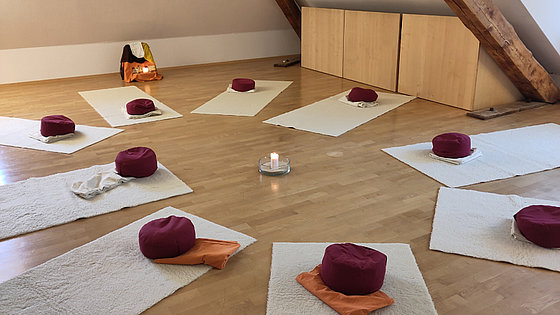 In einem Dachgeschossraum liegen rund um eine brennende Kerze neun einheitliche Teppichläufer mit jeweils einem zusammengelegten Tuch und einem runden Kissen darauf.