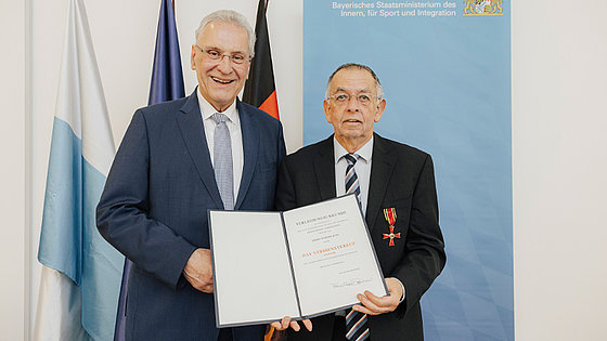 Zwei Herren halten zusammen die Verleihungsurkunde und blicken freundlich in die Kamera: links der Minister, rechts der Geehrte mit dem empfangenen Kreuz an der Brusttasche des Jacketts.