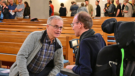Zwei Männer unterhalten sich. Der linke ist der ältere von beiden und trägt Brille, Holzfällerhemd und Strickjacke. Er sitzt am Rand einer Kirchenbank. Der rechte trägt Brille, einen Pullover und einen Schal. Er sitzt in einem Rollstuhl mit Kopfstütze.
