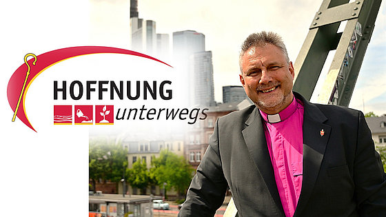 Ein lächelnder Geistlicher in dunklem Sakko und violettem Kollarhemd steht auf einer Brücke vor moderner Stadtkulisse. Links im Bild erscheint das Logo der Initiative »Hoffnung unterwegs«.