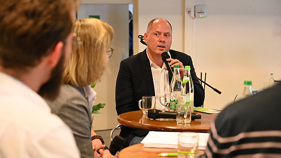 Stefan Weiland, Stirnglatze, kurze Haare, helles Hemd, dunkles Sakko, spricht in ein Mikrofon. Vor ihm sitzen Personen, die ihn ansehen. Auf dem Foto sind Rücken und Hinterköpfe abgebildet. Zwischen Weiland und den Personen stehen zwei runde Tische.