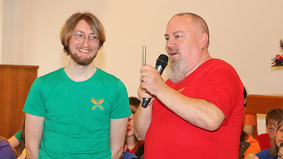Rechts steht ein Mann mit sehr kurzem Haar und langem Bart in rotem T-Shirt mit der rechten Hand ein Mikrofon haltend und in der linken eine Stimmgabel. Links daneben steht ein Mann mit Vollbart und Brille in grünem T-Shirt, der seinen Nachbarn ansieht.