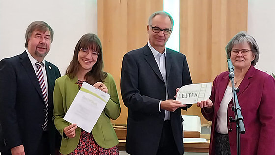 Zwei Frauen und zwei Männer stehen vor dem Abendmahlstisch der EmK-Gemeinde Hamburg-Eppendorf. Superintendentin Irene Kraft (rechts im Bild) überreicht Zertifikat und Plakette zum Abschluss der Gemeindeentwicklung unter dem Titel »Schöpfungsleiter«.