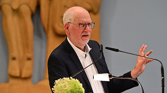 Der Redner, er trägt Brille, dunkles Sakko und offenes weißes Hemd, stützt sich mit dem rechten Ellenbogen auf dem Vortragspult auf. Sein Blick ist seitlich ins Publikum gerichtet. Mit der linken, nach außen geöffneten Hand unterstützt er seine Rede.