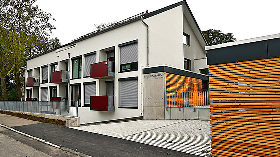 Eine breite Seite mit Fenstern und kleinen Balkonen sowie eine Giebelseite des neuen Wohnheims. Es ist ein zweistöckiges Gebäude plus Dachgeschoss. Direkt vor dem Wohnheim die gepflasterte Abfahrt zur Tiefgarage. Im Vordergrund die Hagstraße.