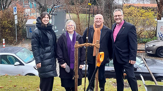 Auf einer Wiese stehen vier Personen nebeneinander: zwei Frauen und zwei Männer. Sie sehen freundlich in die Kamera. Vor ihnen steht der frisch gepflanzte Apfelbaum. Ein Teil eines Spatens ragt ins Bild. Im Hintergrund eine Straße mit Autos.