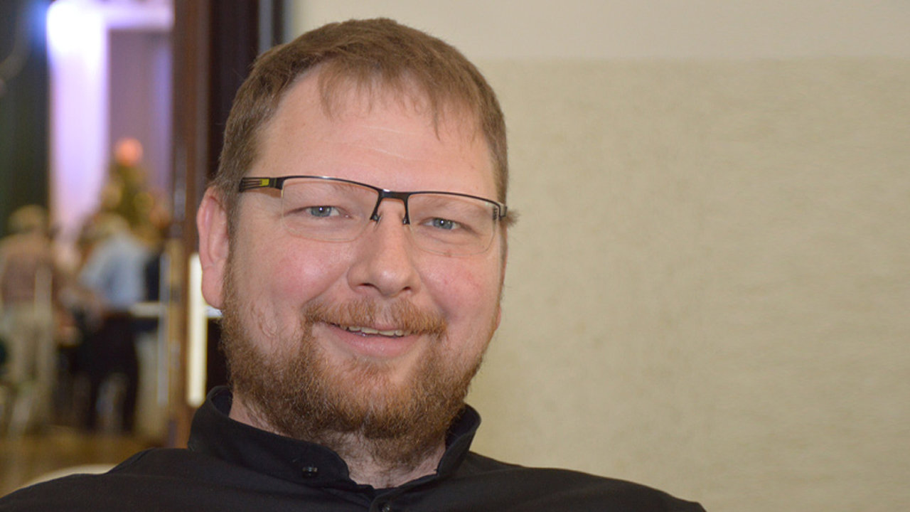 Das Bild zeigt ein Porträt eines Mannes mittleren Alters mit kurzen, hellbraunen Haaren und einem roten Vollbart. Er trägt eine Brille und ein dunkles, langärmeliges Oberteil. Der Hintergrund ist unscharf und zeigt einen hellen Raum mit weiteren Personen.