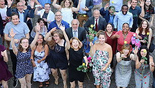 Studierende und Lehrkräfte der THR stehen in einer Gruppe zusammen. Sie sind unterschiedlich gekleidet. Sie lachen und strecken jubelnd die Hände in die Luft. Einige junge Frauen halten eine gelbe Rose in der Hand.