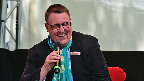Auf einem roten Sofa sitzt ein fröhlich lachender Mann mit Kurzhaarschnitt, Brille und grünem Kirchentagsschal. In der rechten Hand hält er ein Mikrofon und moderiert eine Veranstaltung.