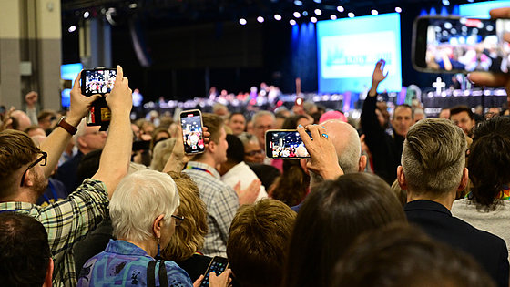 Im Vordergrund drängen sich viele Menschen. Vier von ihnen fotografieren mit ihren Smartphones. Im Hintergrund ist die Bühne in der Halle sichtbar, in der die Generalkonferenz tagt. Über der Bühne: große Anzeigen.