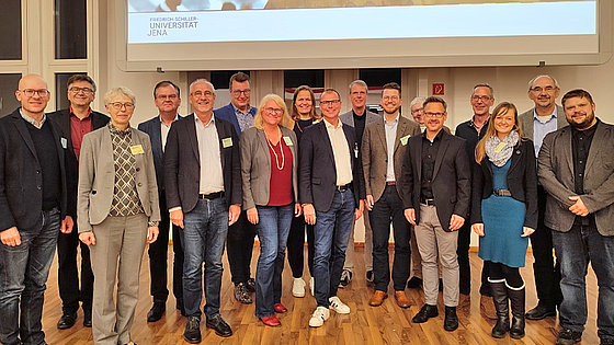 Die siebzehn Personen der Leitung des neuen Studiengangs »Pioneer Ministry« stehen nebeneinander vor der Präsentationsleinwand der Auftaktveranstaltung. Das Bild der Präsentation zeigt ein nach rechts weisendes Holzschild zu einem Hörsaal.