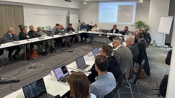 In einem Saal sitzen 21 Menschen an weißen Tischen, die in U-Form aufgestellt sind. Die meisten haben Laptops vor sich stehen. An der Schmalseite des Raums wird ein Text mit der Überschrift »Herzlich verbunden in Vielfalt« an die Wand projiziert.