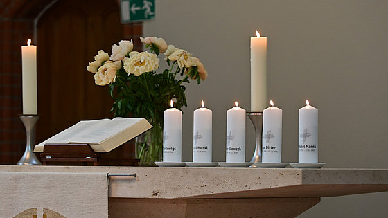 Auf einem Abendmahlstisch aus Stein befinden sich eine aufgeschlagene Bibel, ein Blumenstrauß, zwei hohe Kerzen und fünf mit Namensaufschrift versehene niedrigere Kerzen. Alle Kerzen sind angezündet.